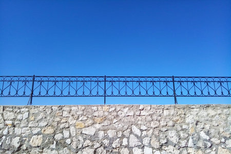 A rough old fence made of gray stone with a figured forged lattice against the background of a blue...