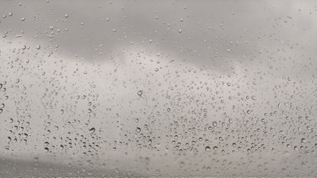 Raindrops on the surface of window panes with gray glowy cloudy background. Black and white horizontal photo. Natural pattern of water droplets. Artificial noise and graininess. Soft focusの写真素材