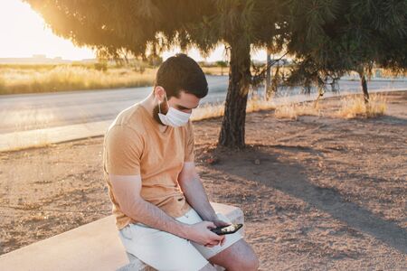 Man in a mask sitting on a bench and texting someone with his smart phone. He is wearing a yellow t-shirt and a white pants.の写真素材
