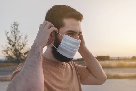 Young man with beard putting on a white face mask to protect himself of coronavirus pandemic. New normality.の写真素材
