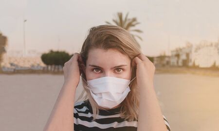 New normal. Young woman portrait putting on a mask and looking at camera with her brown eyes. Blonde girl with a black and white stripes t-shirt. She is in a field enjoying outdoors after covid-19 quarantineの写真素材