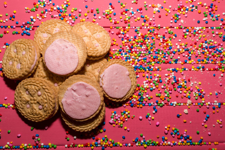 cookies filled with strawberry flavor cream on pink wooden background and colorful sprinkles with space to place textの写真素材