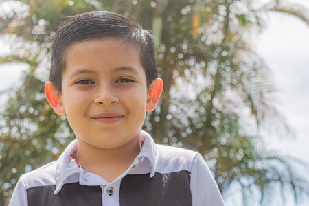 Close-up of a Colombian boy smiling at the camera outdoors, with the background blurred in front of some palm trees on a sunny dayの写真素材