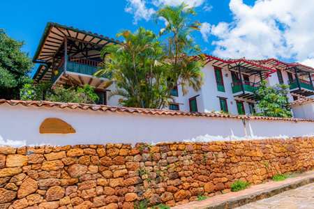 Traditional colonial-style house in Barichara, Colombia, featuring whitewashed walls, red clay roof tiles, wooden balconies, and green-trimmed windows. The foreground showcases a rustic stone wall and cobblestone street, characteristic of this picturesque heritage town. Surrounded by lush tropical plants and under a bright blue sky, this image captures the charm and tranquility of one of Colombia's most beautiful and well-preserved colonial villagesの写真素材