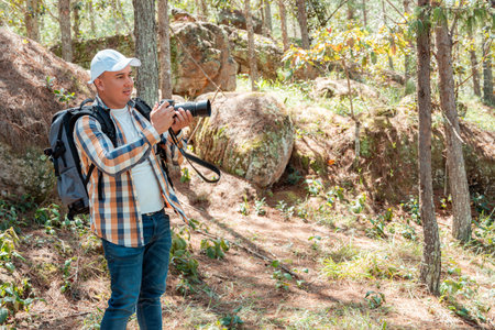 Latin American male photographer with a backpack and camera exploring a sunlit forest. Outdoor...