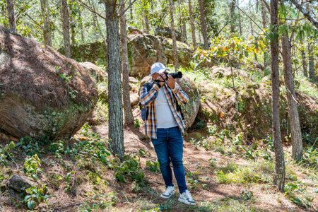Latin American male photographer standing relaxed in forest while taking a photo with DSLR. Natural...