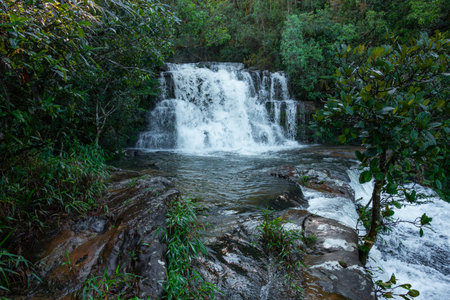Tropical multi-tier waterfall cascading over dark rocks into clear pools, surrounded by lush green...