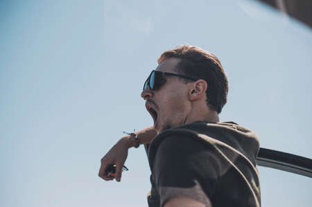 Young old fashioned man in glasses yawns near the car, the clear blue sky on background. Looks tired after long wayの写真素材