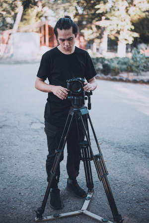 Stylish young operator in black works outdoor with a camera on tripod at the gardenの写真素材