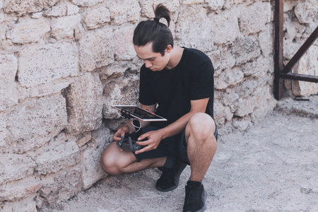 Stylish young drone pilot sits near the wall and uses a remote control. Process of piloting drone outdoors.の写真素材