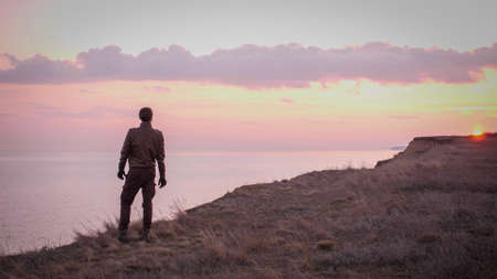 Looking on young biker in leather suit stay on a precipice above sea looking far on majestic sunset with rose clouds.の写真素材