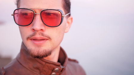 Macho attractive appearance wear leather jacket handsome bearded is troubled about something portrait. His eyes behind red glasses looking upset in sunset light.の写真素材