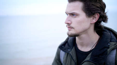 Attractive blue-eyed young man with beard and curly hair lone portrait on a windy cliff above dark sea. Looking worried, He look up on troubled water and cloudy sky with a hope at late autumnの写真素材