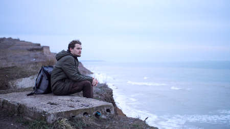 Sad lonely young man, with backpack, sits on a cement block at the edge of cliff above storm sea, and looks in a far lost in thought. Mist adds a moody atmosphere in late autumn cloudy dayの写真素材