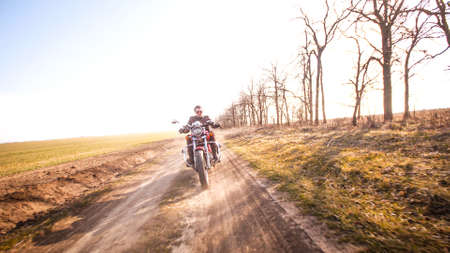 Young curly biker with red glasses drive ride on red chopper through the desert dirt road in late autumn sunset lightの写真素材