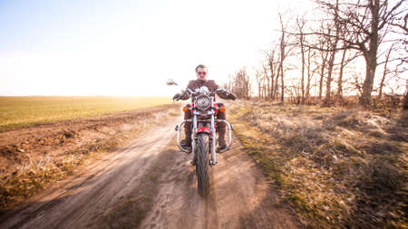 Young curly biker with red glasses drive ride on red chopper through the desert dirt road in late autumn sunset lightの写真素材