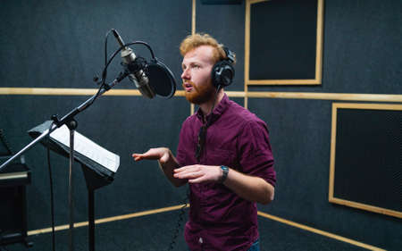 Expressive bearded man with curly ginger hair in headphones at recording studio stay opposite a microphone and performs like voice actorの写真素材