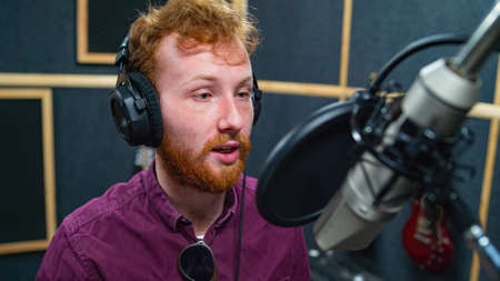 Expressive bearded man with curly ginger hair in headphones at recording studio stay opposite a microphone and performs like voice actorの写真素材