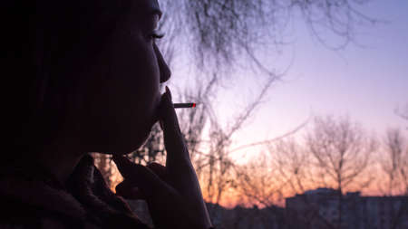 Pretty young woman with curly hair and long nails makes deep breath before smoke a joint with marijuana looking on sunset in open window. Depress atmosphere of teen decay after using narcotic.の写真素材