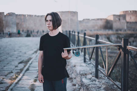 Young longhaired man in sport outfit stay with skateboard on the bridge with ancient castle backgroundの写真素材