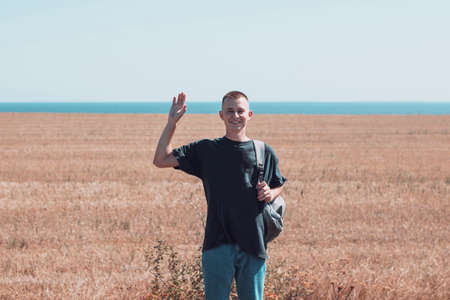 Happy young man with short hair smile and shake a hand to a camera on a field in summer sunny weather with a sea on backgroundの写真素材