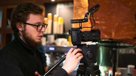 Professional operator uses a camera on tripod to set a frame in the cafe. Behind bearded young operator in glasses scenic dot lights and reflectors.の写真素材