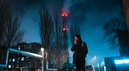 Shoot of a smoking modern man in hooded coat and lighted with neon color in the night industrial zone on background.の写真素材