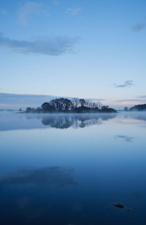 Reflection of trees in the calm water of a lake at sunriseの素材