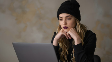 Portrait of a young woman in a black hat sitting at a table with a laptopの素材