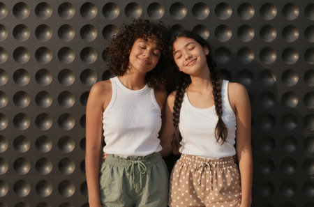 Two teenage girls in white T-shirts and green pants on the background of a metal wallの素材