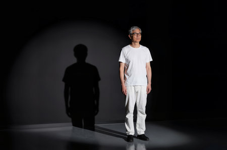 Portrait of an Asian senior man wearing white t-shirt and jeans standing in a dark roomの素材