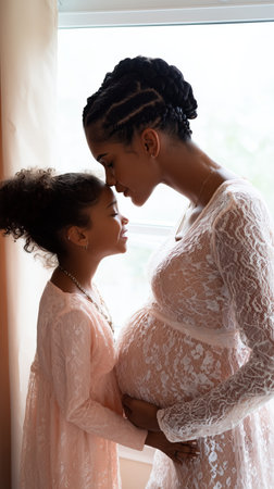 A pregnant mother and her young daughter gently touch foreheads, exchanging smiles. They stand near a window, their soft dresses creating a cozy atmosphere.の素材