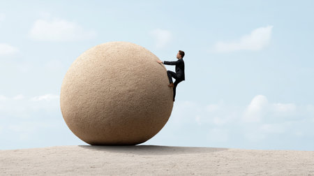 A man dressed in a suit climbs a large round rock in a vast landscape. The scene captures the effort and determination of the man as he scales the smooth surface under a bright sky.の素材