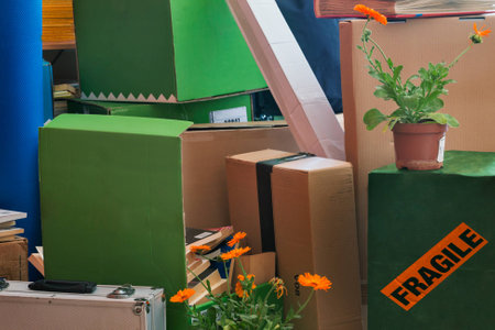 A stack of different size boxes ready to move. Several books appear between the boxes, in the foreground there is a metal case and a potted flower, in the background a photo album and another potted flower.A sticker on a box saysの写真素材
