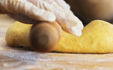 A rolling pin is used to flatten the dough  ,in the foreground  one hand with white latex glove , on the wooden board a bit of white flourの写真素材