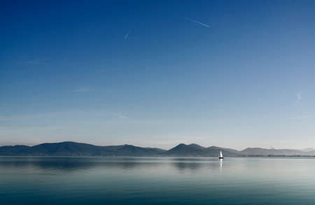 One sail boat in the calm waters of Lake Trasimeno, Italy, in the background small townsの写真素材