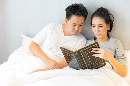 Young Asian Couple laid in a bed reading a book in bedroom of contemporary house for modern lifestyle conceptの写真素材