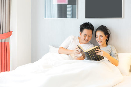 Young Asian Couple laid in a bed reading a book in bedroom of contemporary house for modern lifestyle conceptの写真素材