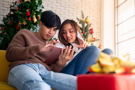 Young Adult teenager couple using tablet on sofa in living room for online shopping with gift box with Christmas tree in backgroundの写真素材