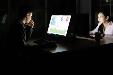 Young adult asian businessman and woman working late at night in their office with desktop computer...