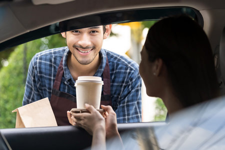 Waiter giving hot coffee cup with disposable tray and bakery bag through car window to customer at...