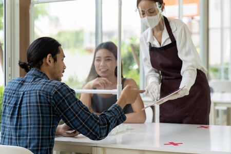 Asian waitress wear face mask and face shield using tablet to show restaurant electronic menu and recommend to customer. New normal technology and lifestyle food and dining concept.の写真素材