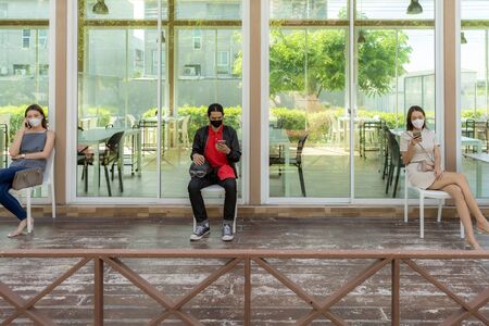 Asian customers wear face mask using mobile phone while sit in waitting social distance queue in front of restaurant with free copyspace for editing. New normal restaurant concept.の写真素材