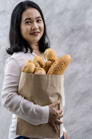 Portrait of asian smile woman housewife holding variety bread in disposable paper bag on grey vintage loft background. Bakery food and drink grocery and domestic life lifestyle concept for delivery.の写真素材
