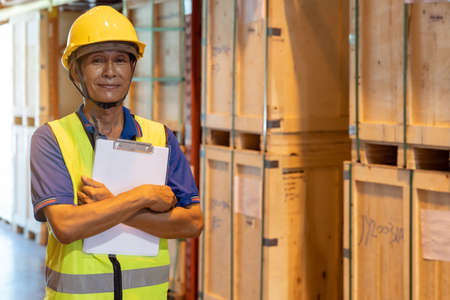 Portrait of Asian warehouse worker with clipboard job cross arm in large distribution warehouse background. Reopening business warehouse inventory and logistic concept.の写真素材