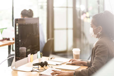 Mixed race of African black and asian businesswoman office worker wear protective face mask working in new normal office with social distance between table to prevent coronavirus COVID-19 spreading.の写真素材
