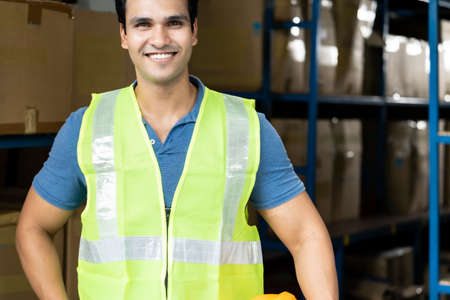 Portrait of Indian asian warehouse worker with safety vest stand and hold yellow helmet in warehouse distribution center environment.の写真素材