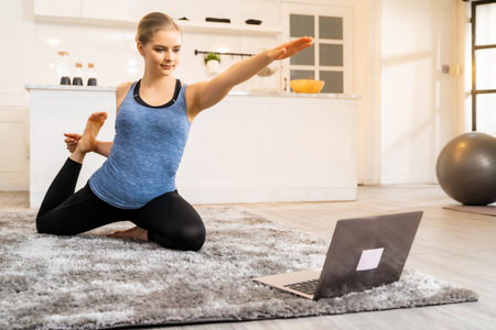 White caucasian fit sporty woman doing yoga king pigeon pose while working at home with laptop in...