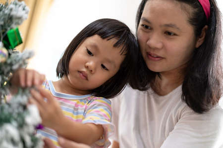 Little asian girl with mom decorating a Christmas tree in her living room with christmas ornament preparing for season greeting together. Multigenerational Family engage and happiness concept.の写真素材