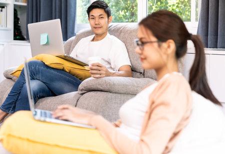 Asian working man use computer notebook laptop work from home in living room with social distance to his couple in background while city lockdown. Work at home concept.の写真素材